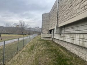 This image shows a pathway running alongside a chain-link fence next to a large retaining wall or noise barrier. The wall appears to be part of highway infrastructure, with a sloped embankment and an excavator working near the top.