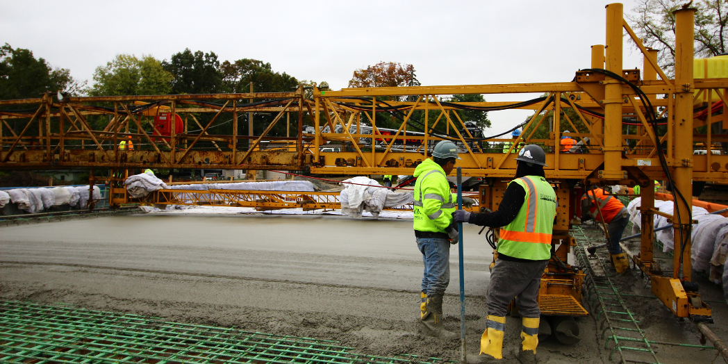 This image shows a bridge construction site where workers in high-visibility gear and hard hats are actively engaged in pouring and smoothing concrete over a reinforced steel rebar structure.