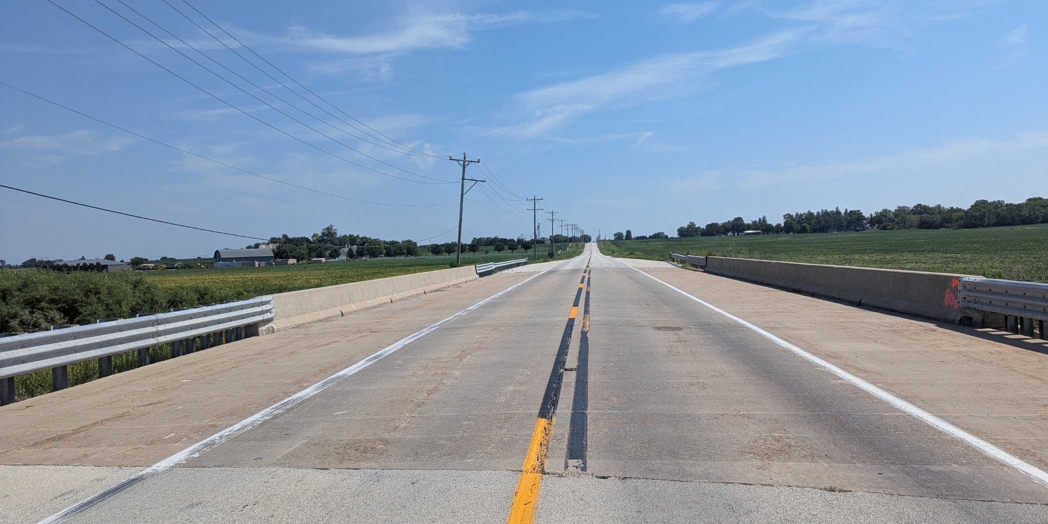 View across an existing County Line Road bridge with concrete barriers, metal guardrails, and farmland on both sides.