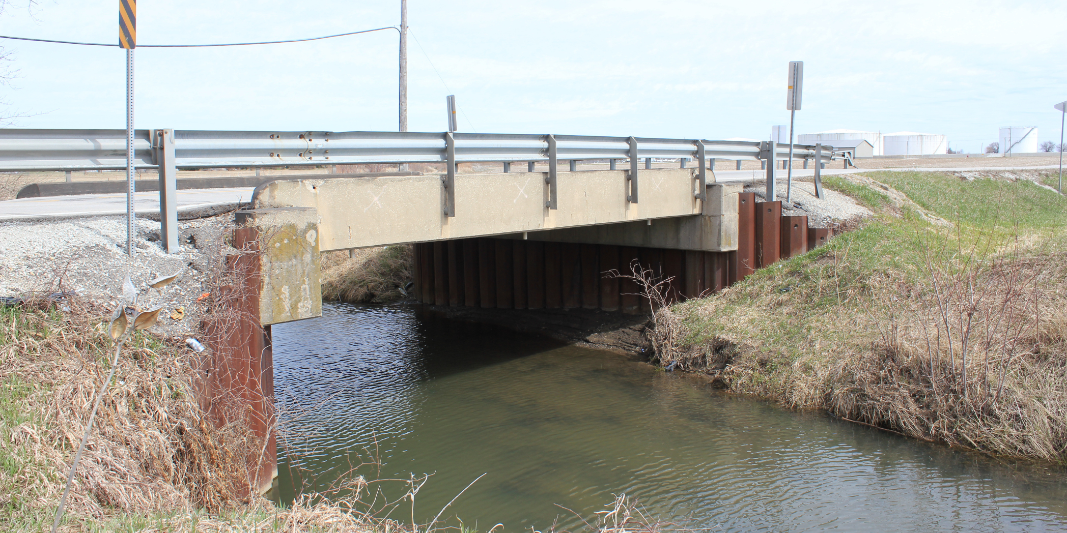 Side view of an existing County Line Road bridge over a waterway, with concrete supports, metal guardrails, and grassy banks.