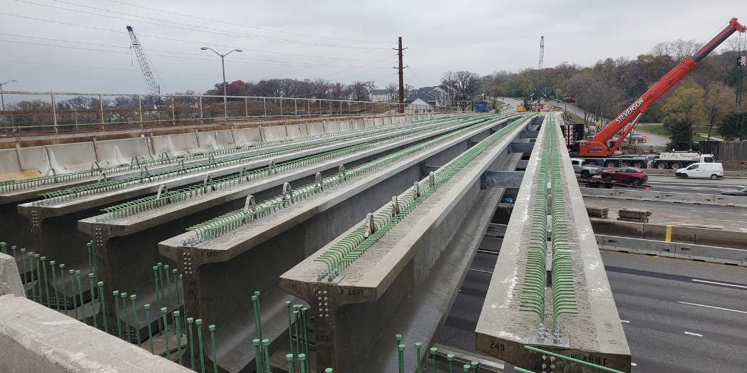 Parallel concrete bridge girders with green reinforcing bars span I-294, with cranes and highway traffic visible below.