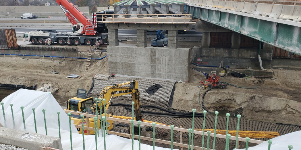 Concrete bridge pier beside Flagg Creek, with an excavator, crane, and erosion protection mats during Plainfield Road bridge replacement.