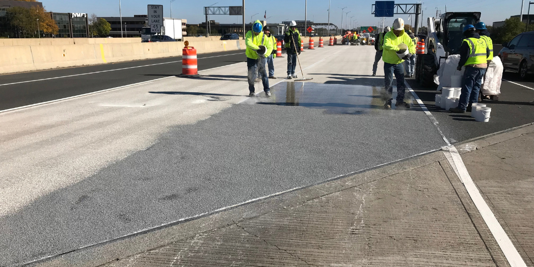 Workers spread material across an I-88 bridge deck beside construction equipment and buckets in a closed lane.