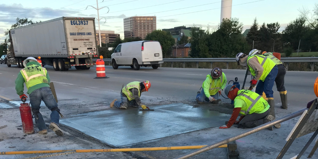 Workers finish a rectangular concrete pavement patch on I-88 while vehicles travel in the adjacent lane.