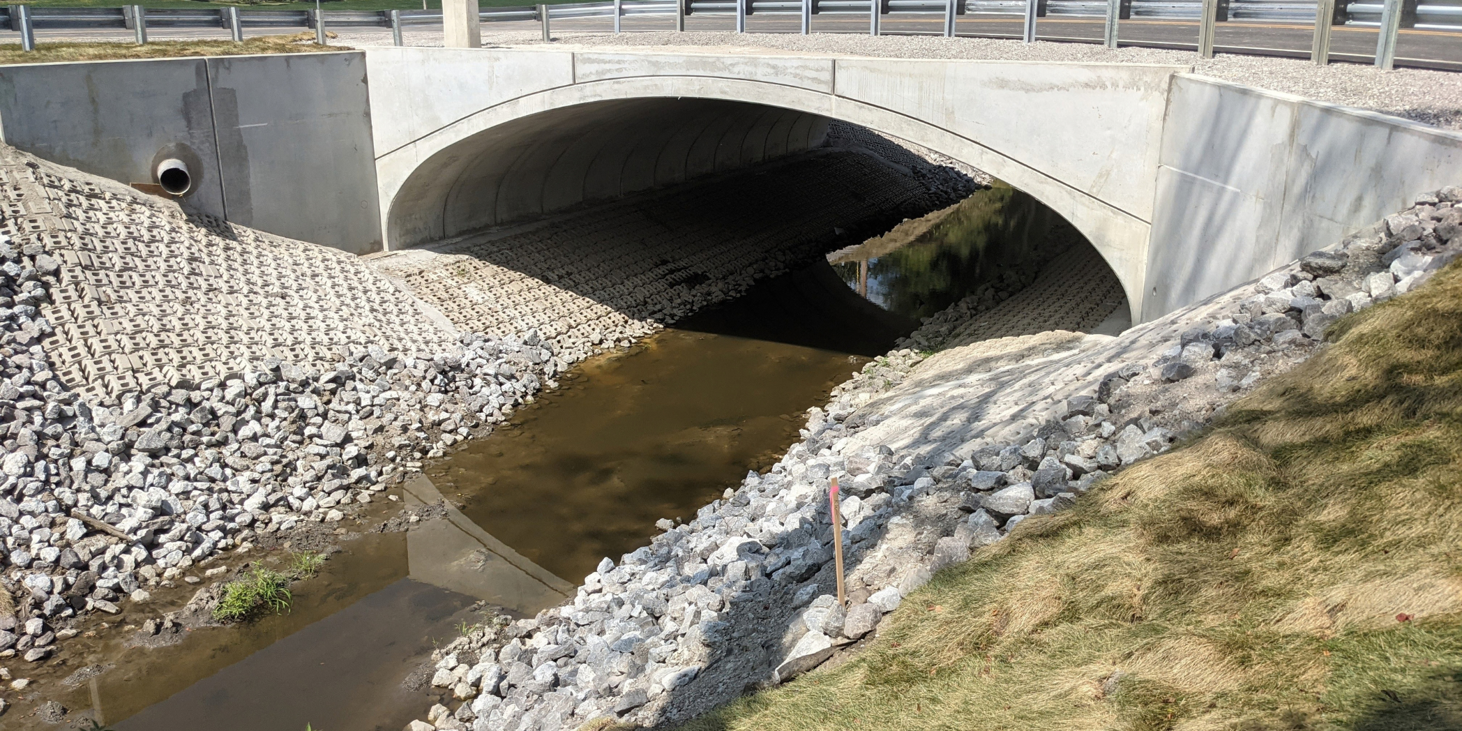 Completed concrete arch at Bridge No. 65 spans a stream, with concrete wingwalls, rock slope protection, and roadway guardrail above.