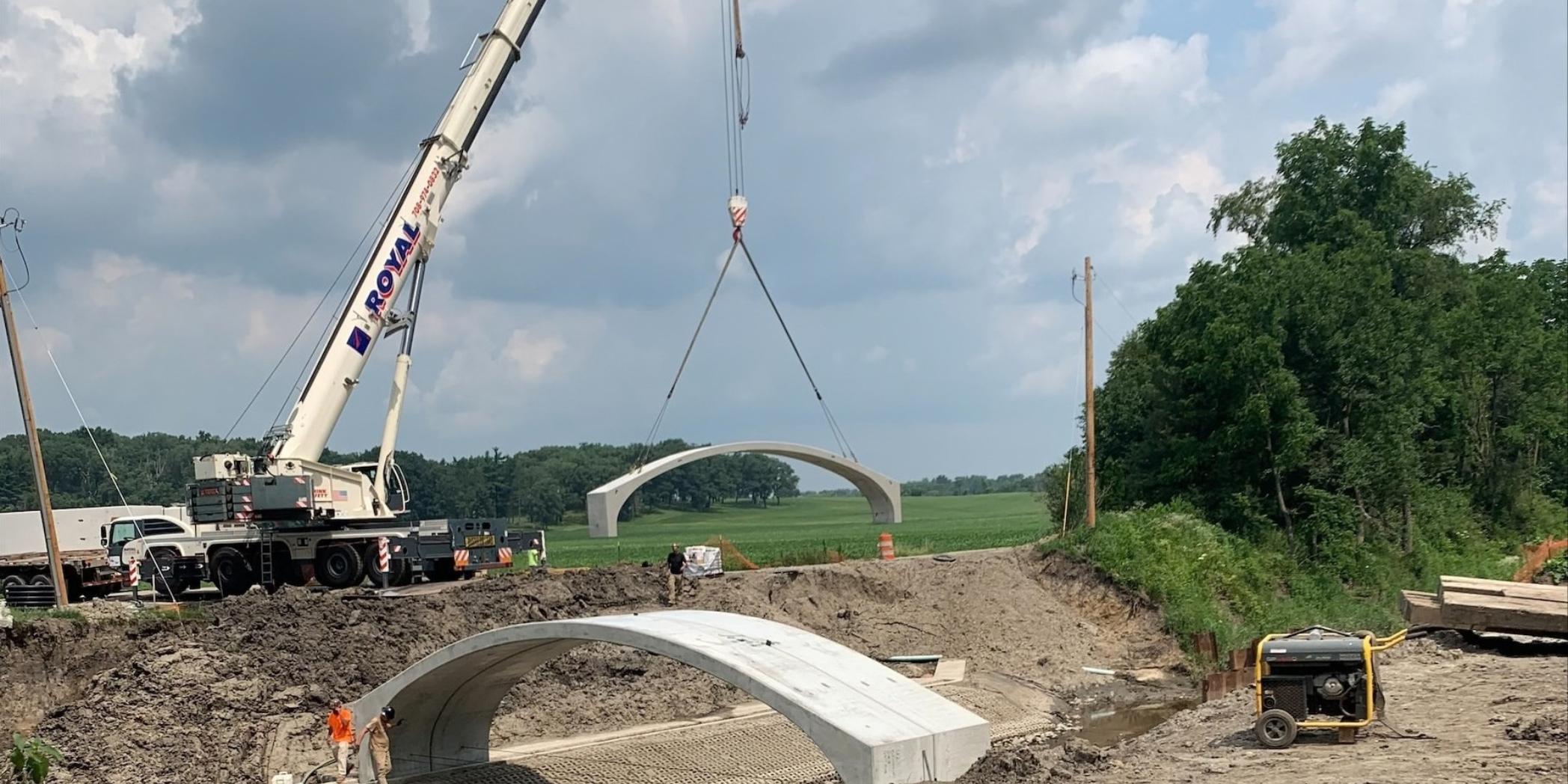 A crane lifts a precast concrete arch section into place at Bridge No. 65, with workers beside previously installed sections.