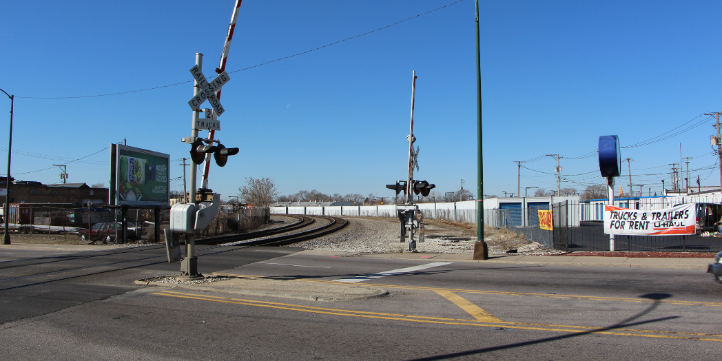 Two railroad tracks curve across Central Avenue at street level, with raised crossing gates, warning lights, and railroad crossing signs.