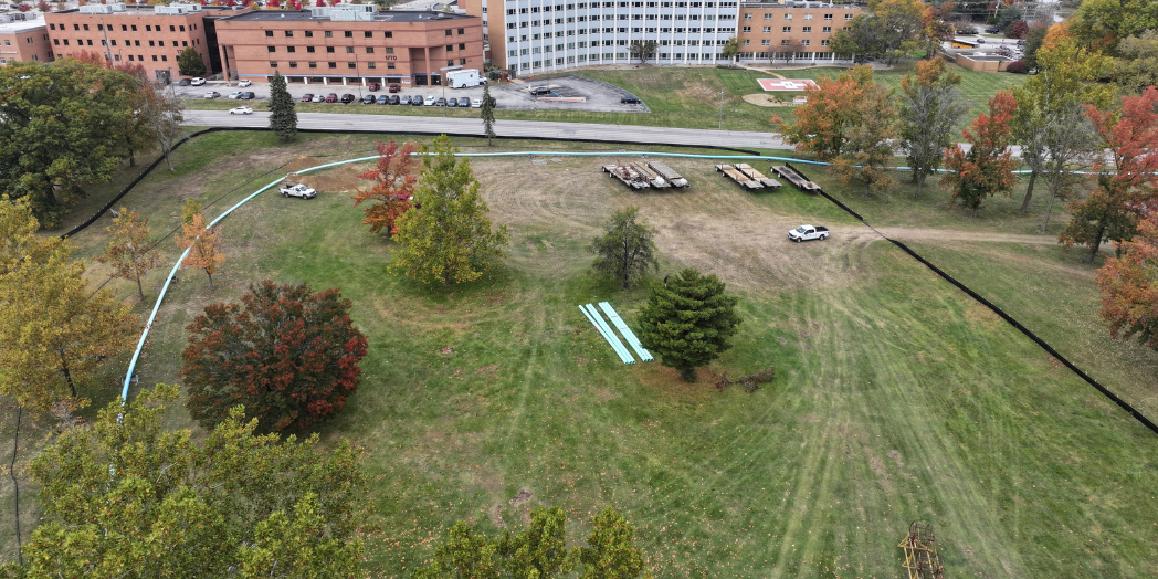 Aerial view of a long force main pipe across a grassy area near buildings, roads, and trees with fall foliage.