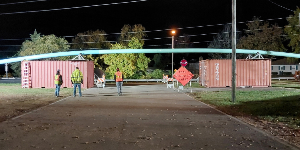 Workers monitor a force main pipe elevated above a barricaded street on supports atop shipping containers at night.