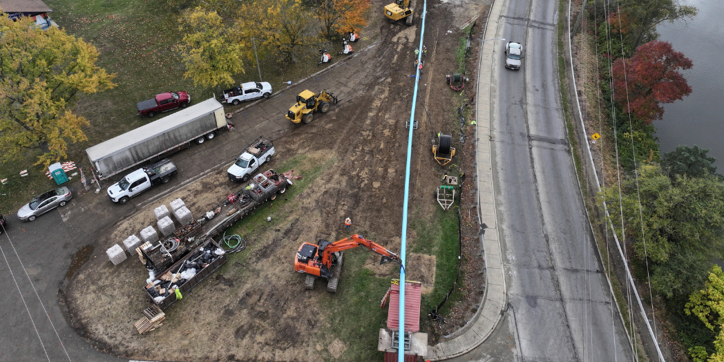 Aerial view of a force main pipe beside a lakeside road, with construction crews, excavators, and work vehicles nearby.
