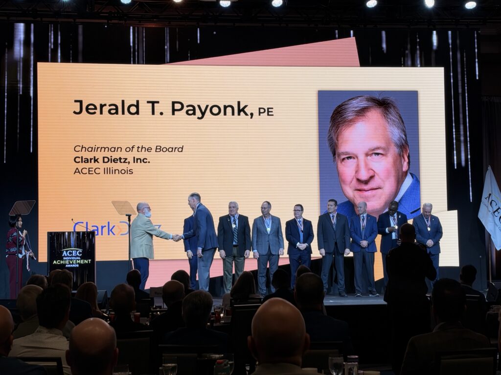 Ceremony screen displays Jerald T. Payonk, PE, Chairman of the Board at Clark Dietz, with his portrait as participants stand on stage.