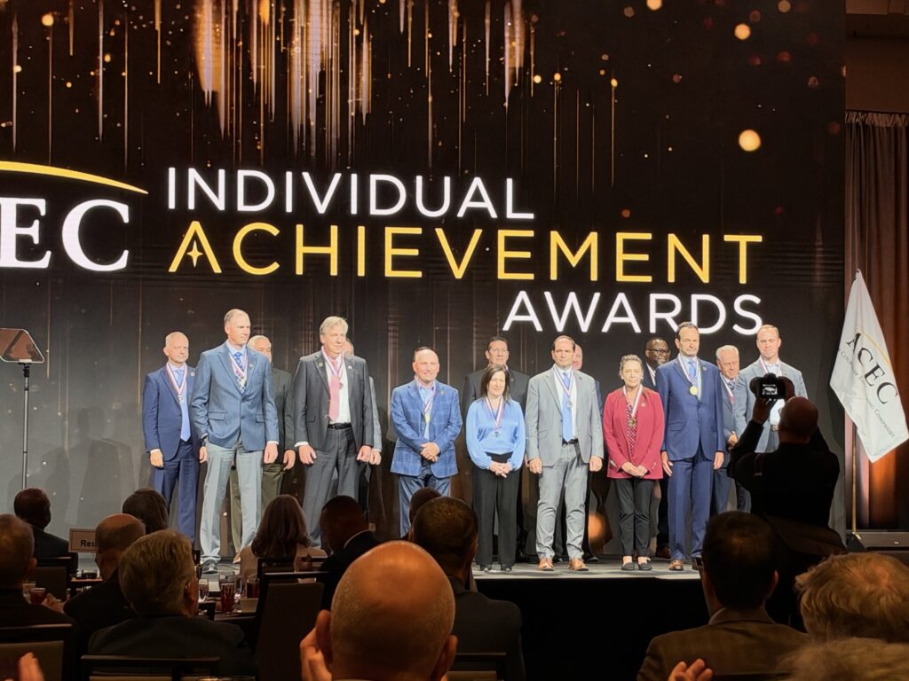 College of Fellows inductees wearing medals pose on stage beneath an ACEC Individual Achievement Awards display.